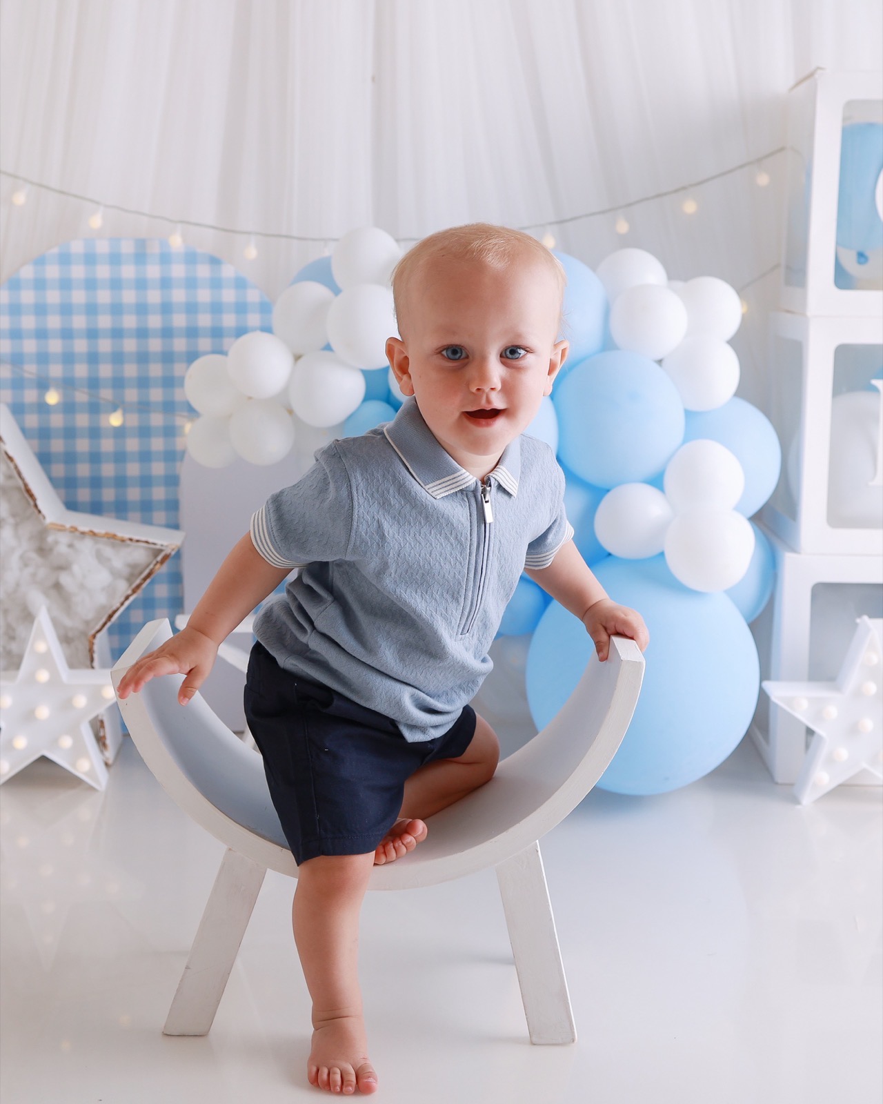 Blonde toddler boy standing on a curved white bench prop, grey zip-up polo and navy shorts, blue and white balloon backdrop