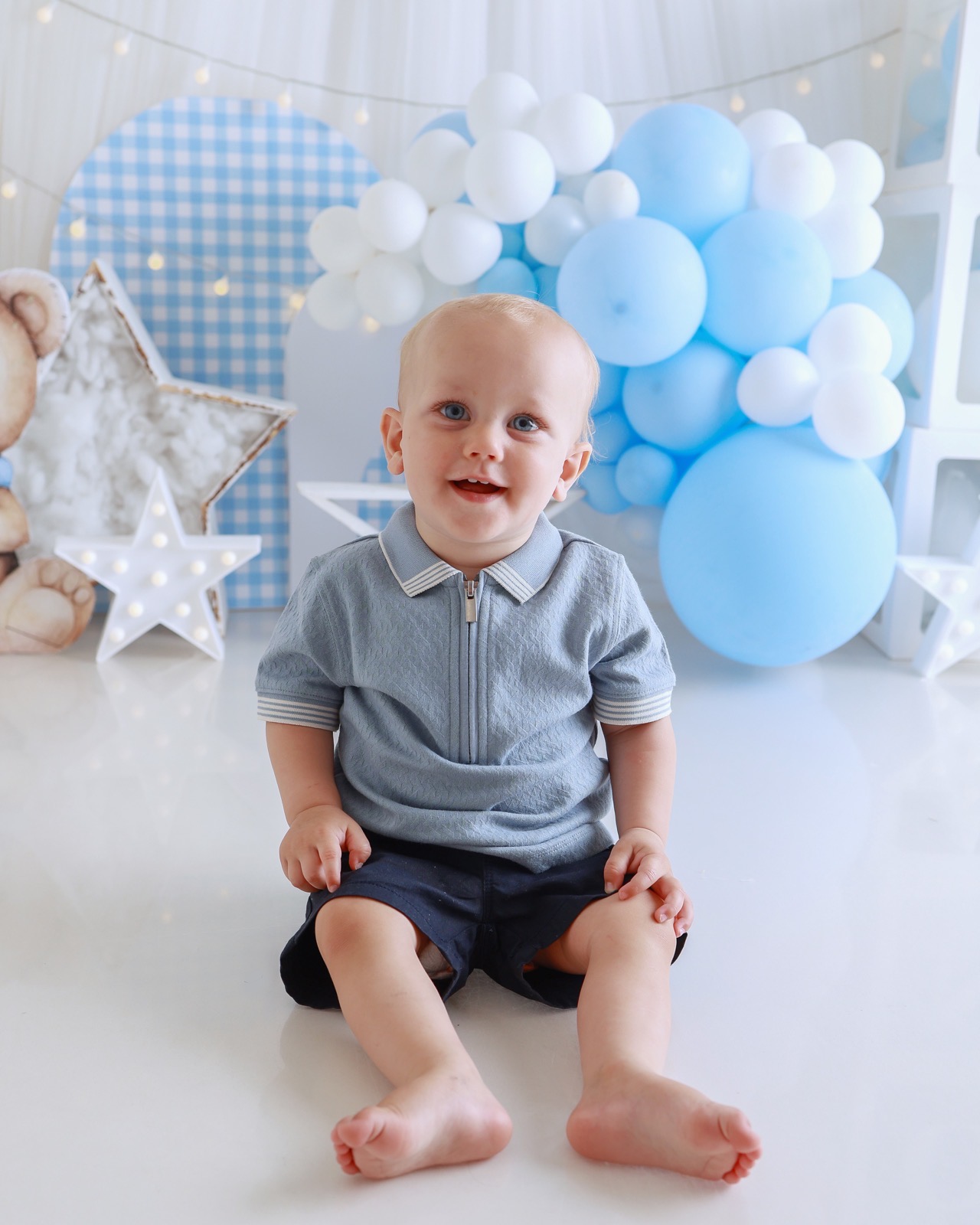 Blonde toddler boy sitting on the floor smiling, grey zip-up polo and navy shorts, star-themed balloon backdrop