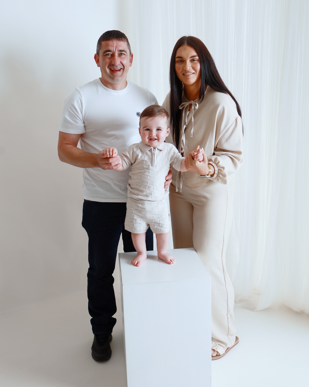 Family of three, toddler boy standing on a white plinth holding hands with his mum and dad