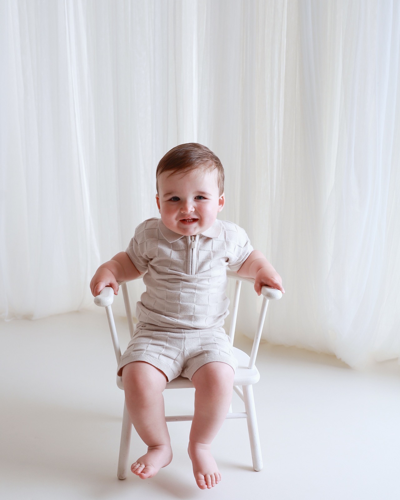 Toddler boy in a beige knitted outfit sitting on a small white chair