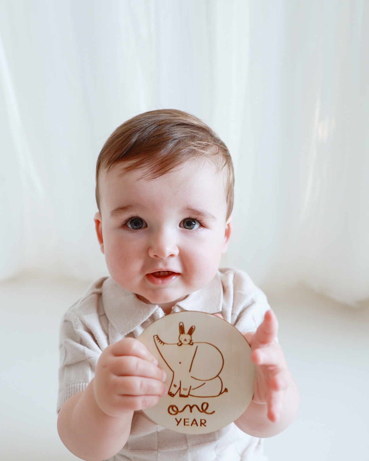 Close-up of a toddler boy holding a wooden 'one year' milestone disc with a bunny and elephant illustration