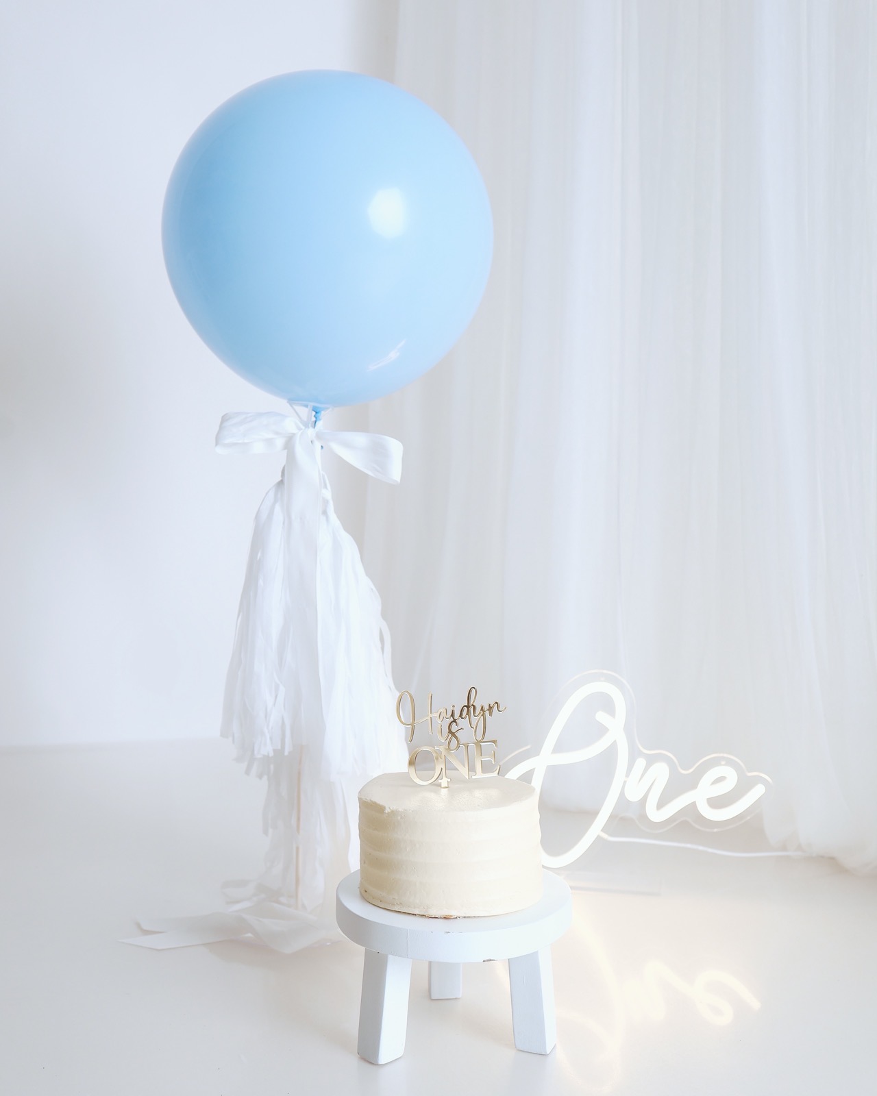 A plain cream birthday cake with a personalised topper, a large blue balloon and a lit-up 'One' sign