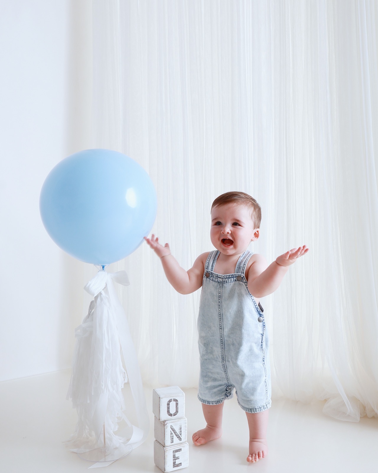 Toddler boy in light-wash denim dungarees reaching for a large blue balloon, upset expression, 'ONE' blocks nearby