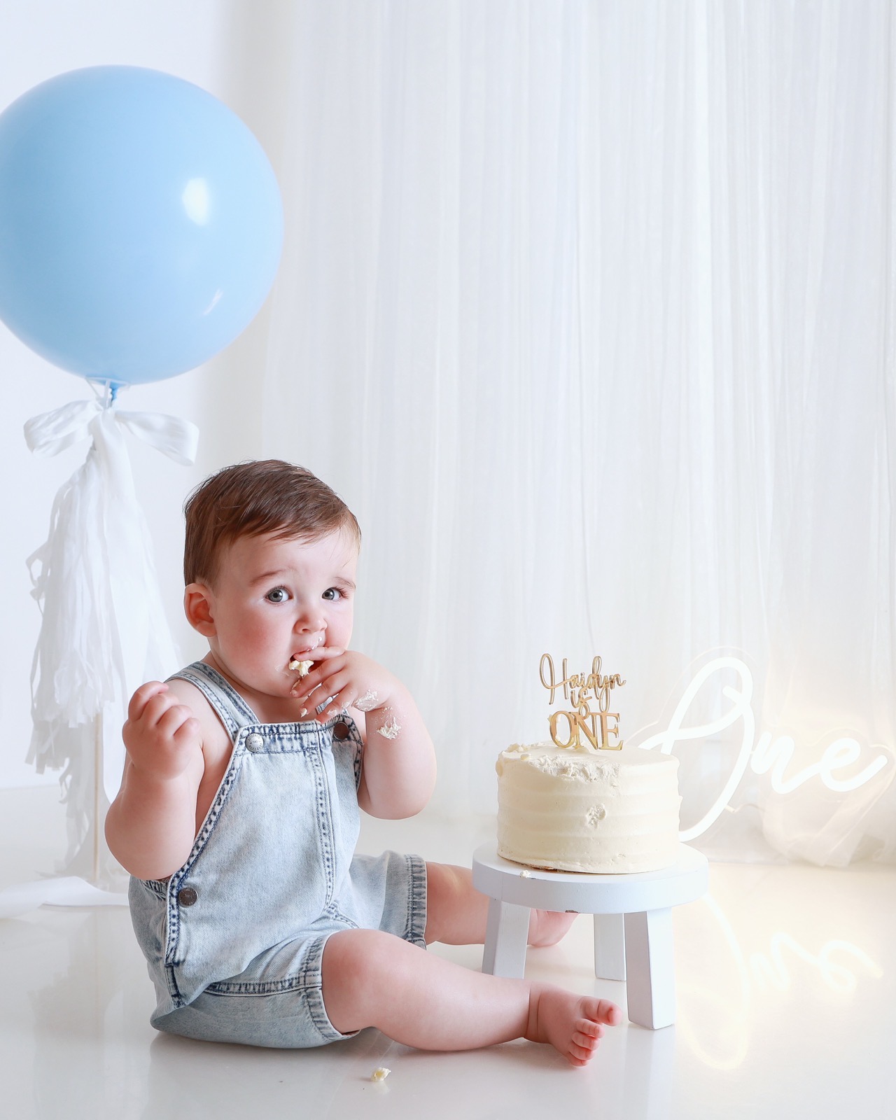 Toddler boy sitting and eating cake with his fingers, blue balloon beside him, lit-up 'One' sign behind