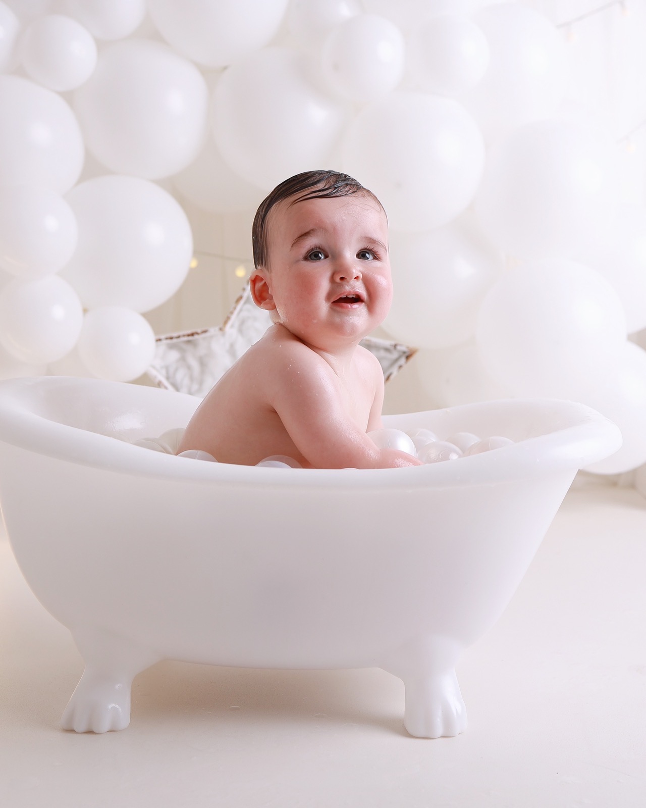 Toddler boy with damp hair sitting in a white clawfoot bath tub filled with white bath balls, white balloon backdrop