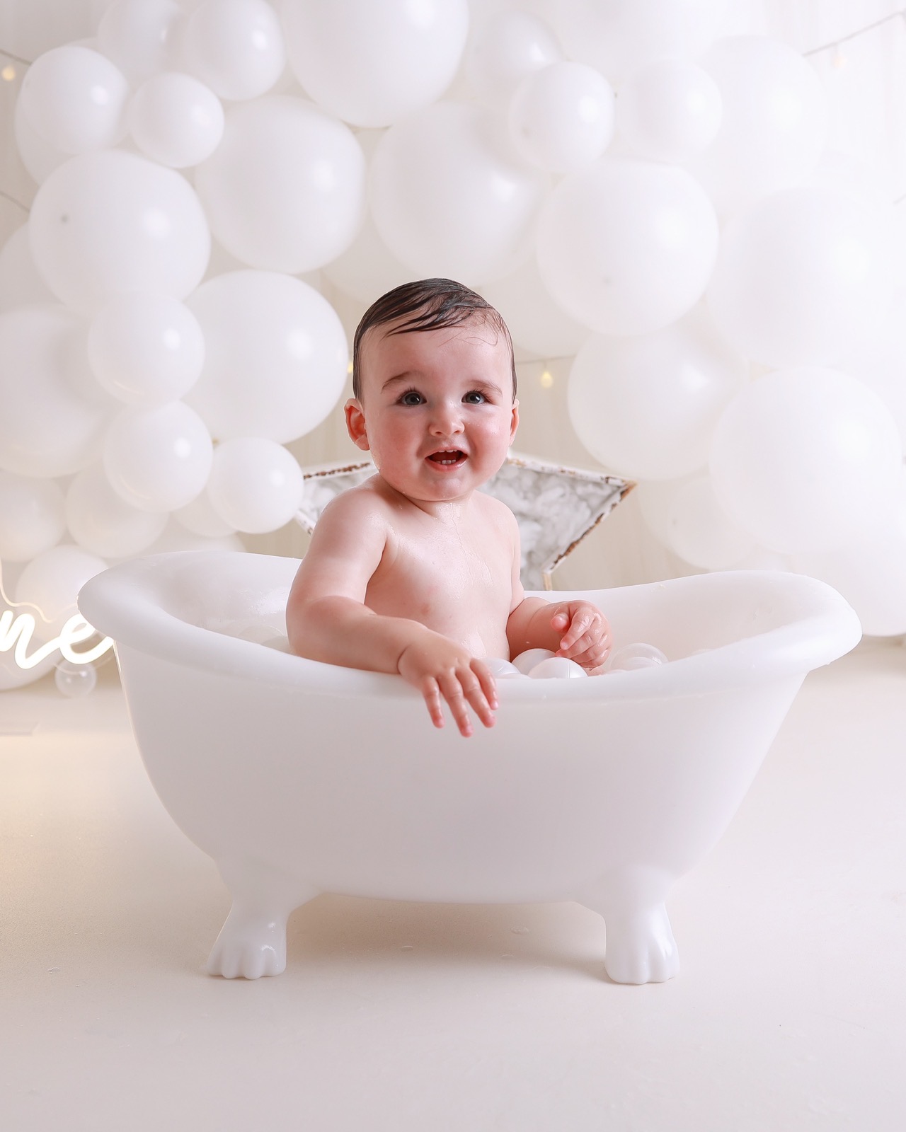 Toddler boy smiling in a white bath tub surrounded by white bath balls, a marbled star prop behind him