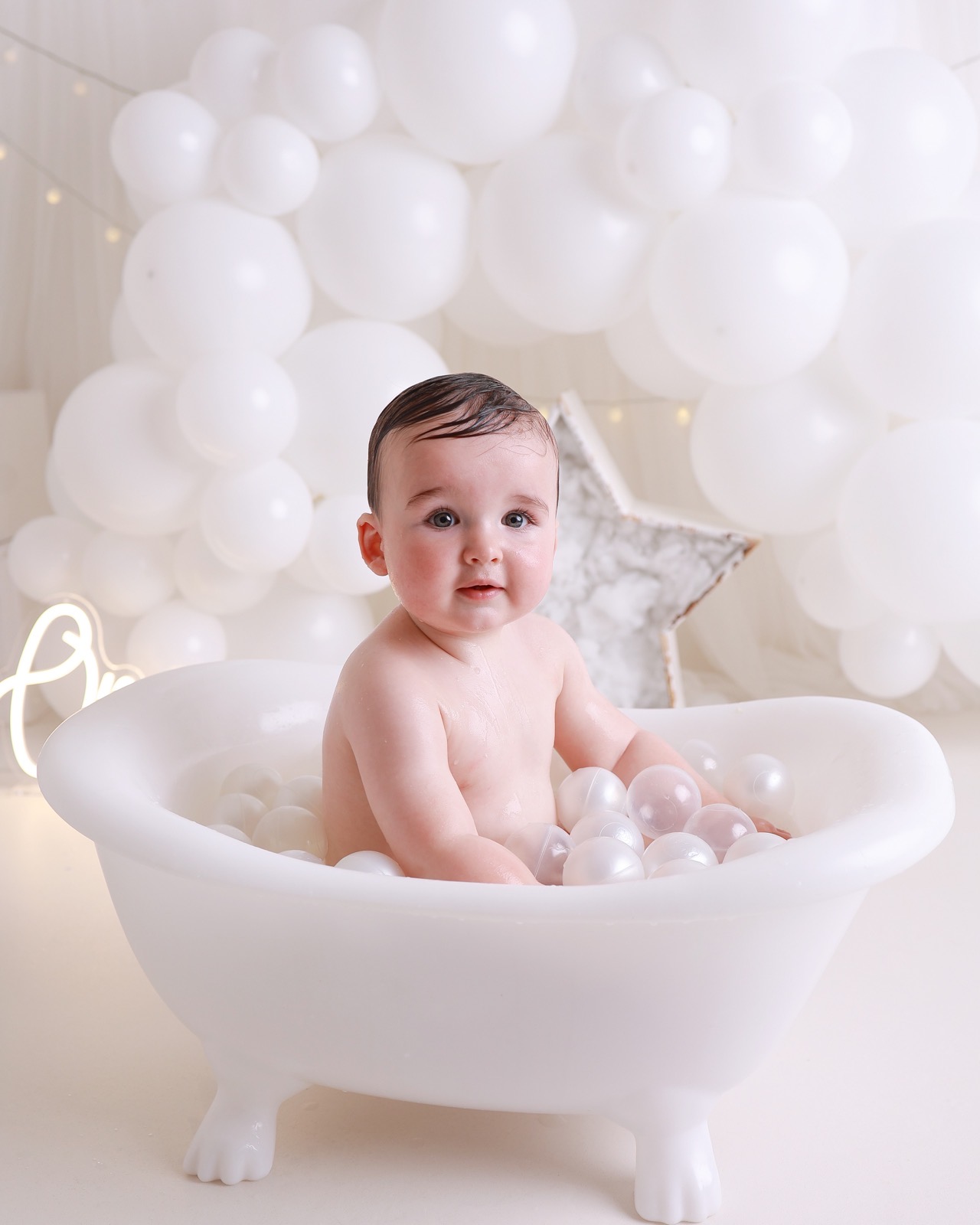 Toddler boy sitting in a white bath tub full of pearl-white bath balls, an illuminated 'One' sign glowing behind