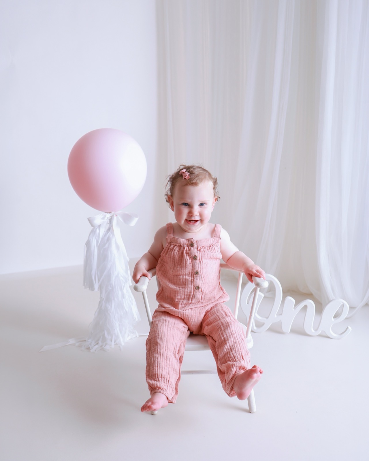 Baby girl in a dusty pink jumpsuit sitting on a white chair beside a pink balloon and a wooden 'One' script sign