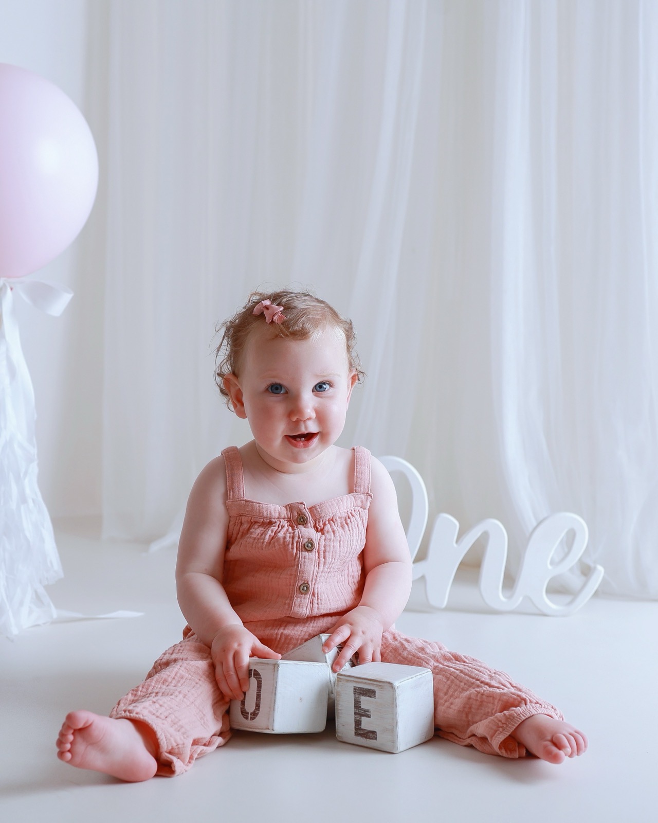 Baby girl sitting on the floor with wooden 'ONE' blocks, pink balloon and 'One' script sign behind
