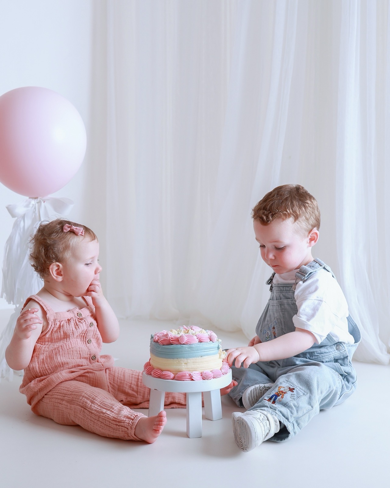 Twin siblings sitting together with a pink, blue and cream birthday cake between them, brother holding a spoon