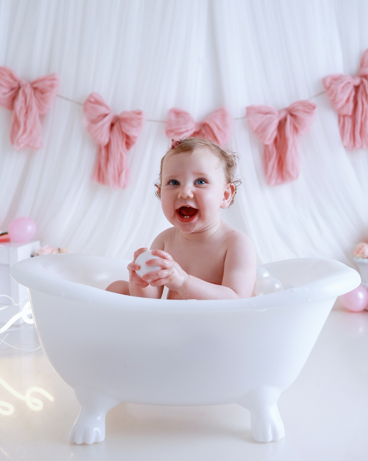 Baby girl laughing in a white bath tub, pink yarn bow bunting behind, holding a bath ball