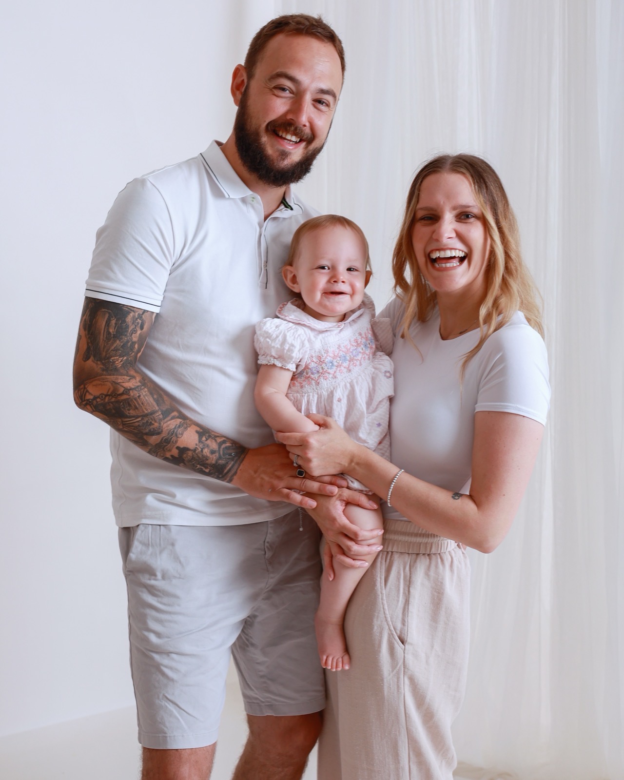 Family of three, dad with a tattoo sleeve and mum in beige loungewear each holding their baby daughter in a floral dress