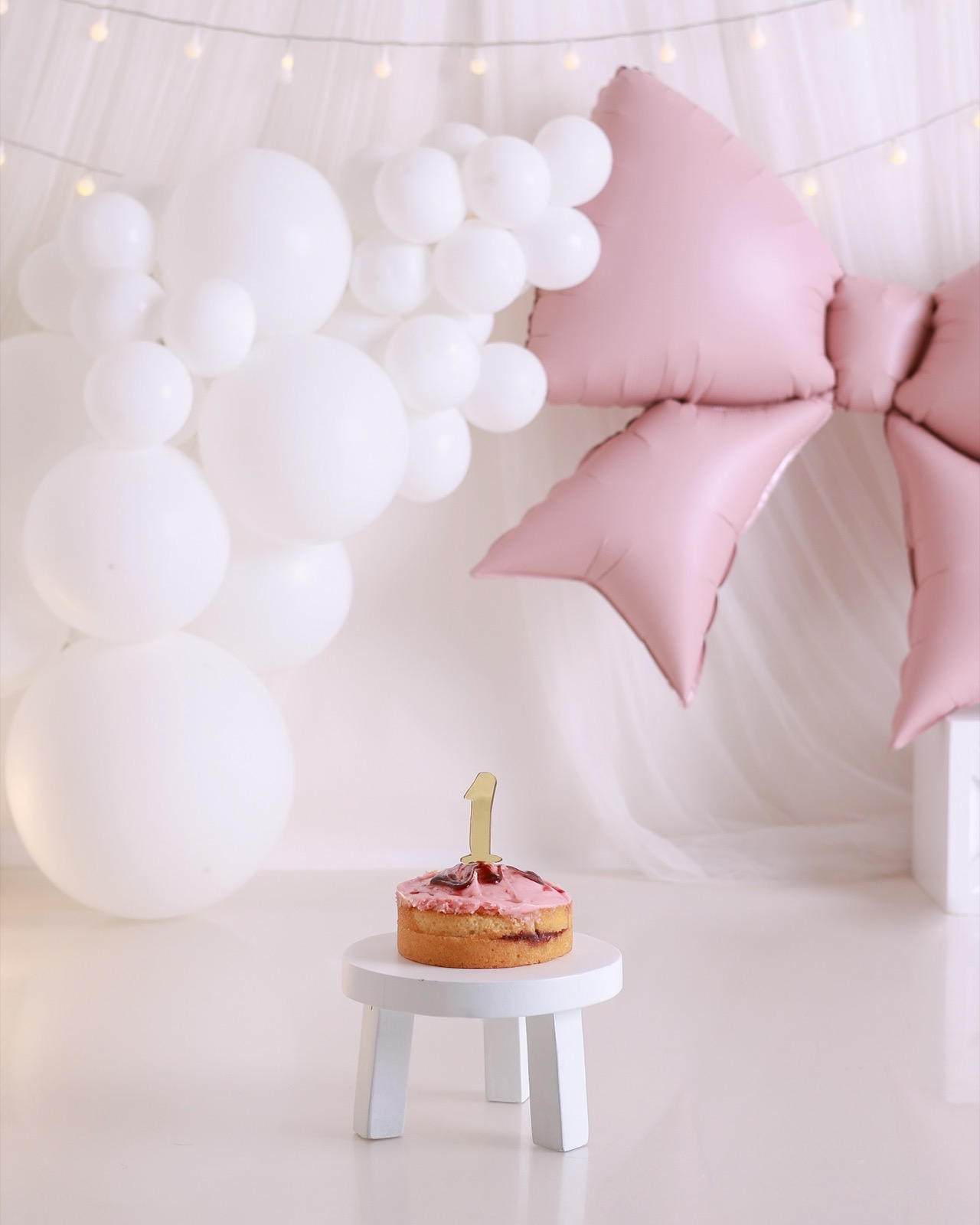 A small round sponge cake with pink icing, cherries and a gold '1' topper, pink bow balloon and white balloon garland behind