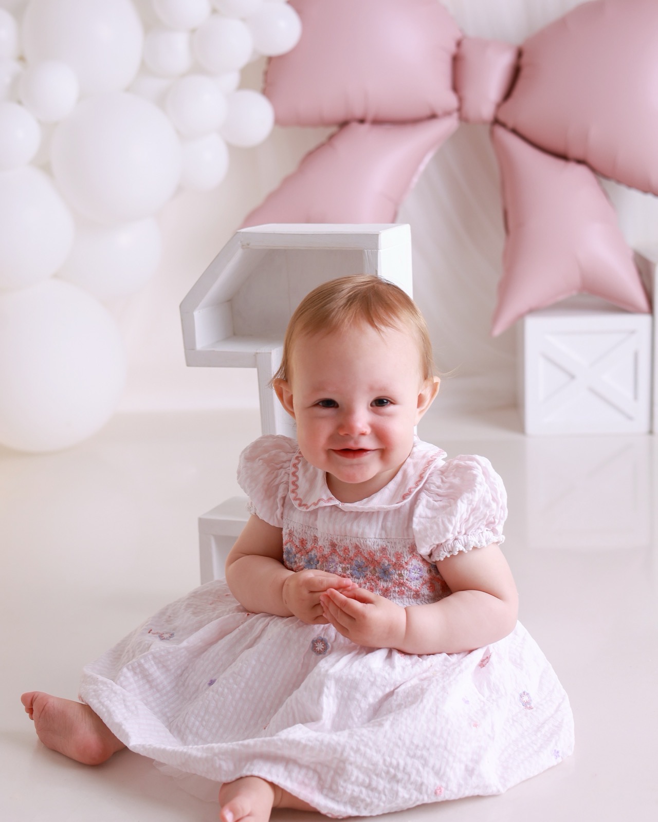 Baby girl in a pale pink smocked dress sitting beside a large white wooden '1' prop and a pink foil bow balloon