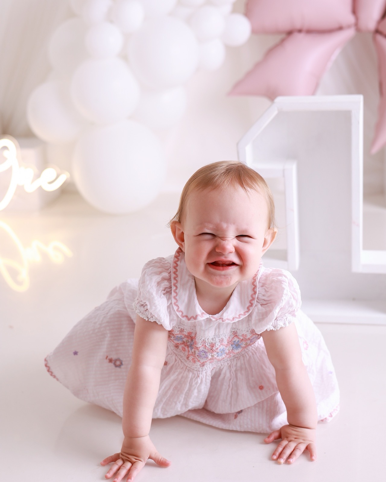 Baby girl in a pink floral dress crawling toward the camera with a scrunched-up expression, '1' prop and bow balloon behind