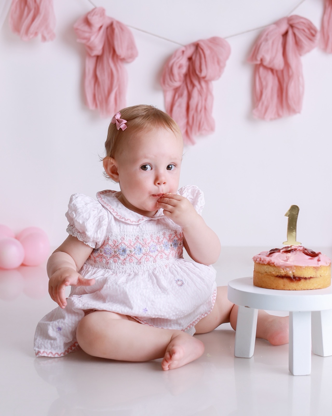 Baby girl tasting cake with her fingers, small round pink-iced sponge cake with a gold '1' topper, pink tassel garland behind
