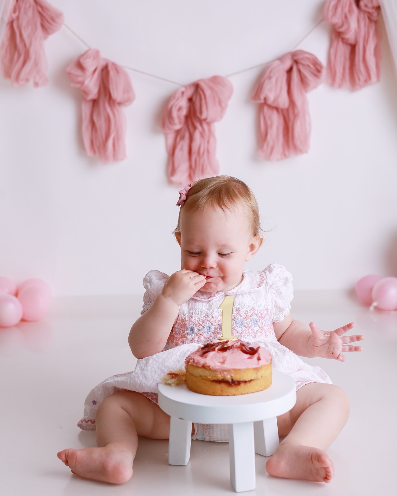 Baby girl savouring a bite of cake with her eyes closed, pink tassel garland backdrop