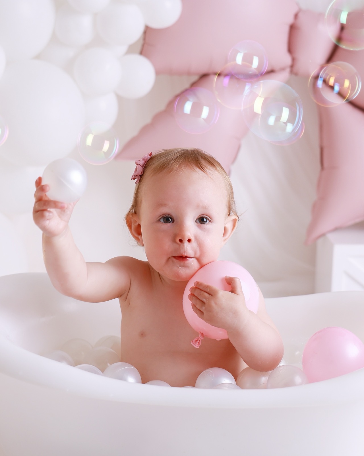 Baby girl in a white bath tub holding a bath ball and a pink balloon, soap bubbles floating around her