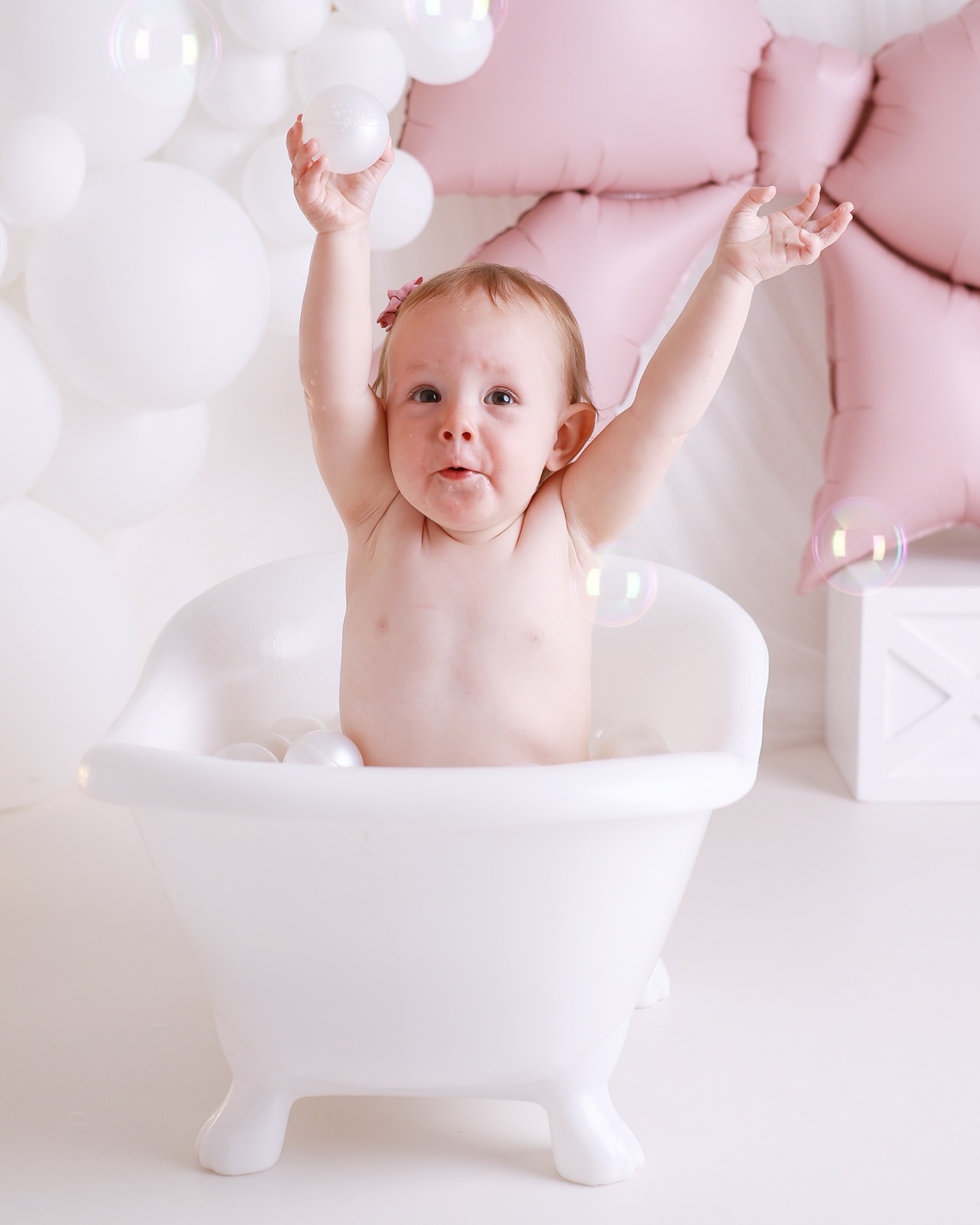 Baby girl in a white bath tub with both arms raised, holding a bath ball, pink bow balloon backdrop