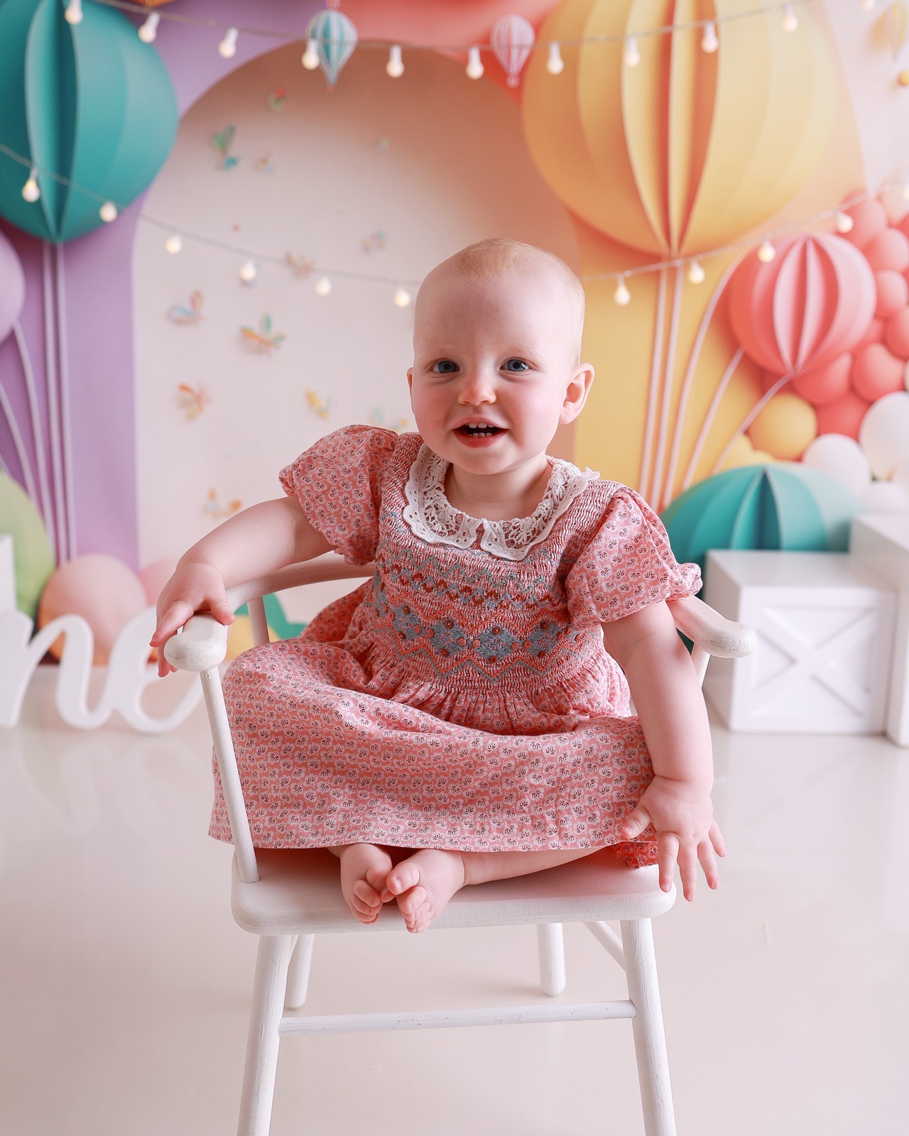 Baby girl in a pink floral dress sitting on a white chair, colourful hot-air-balloon-themed backdrop