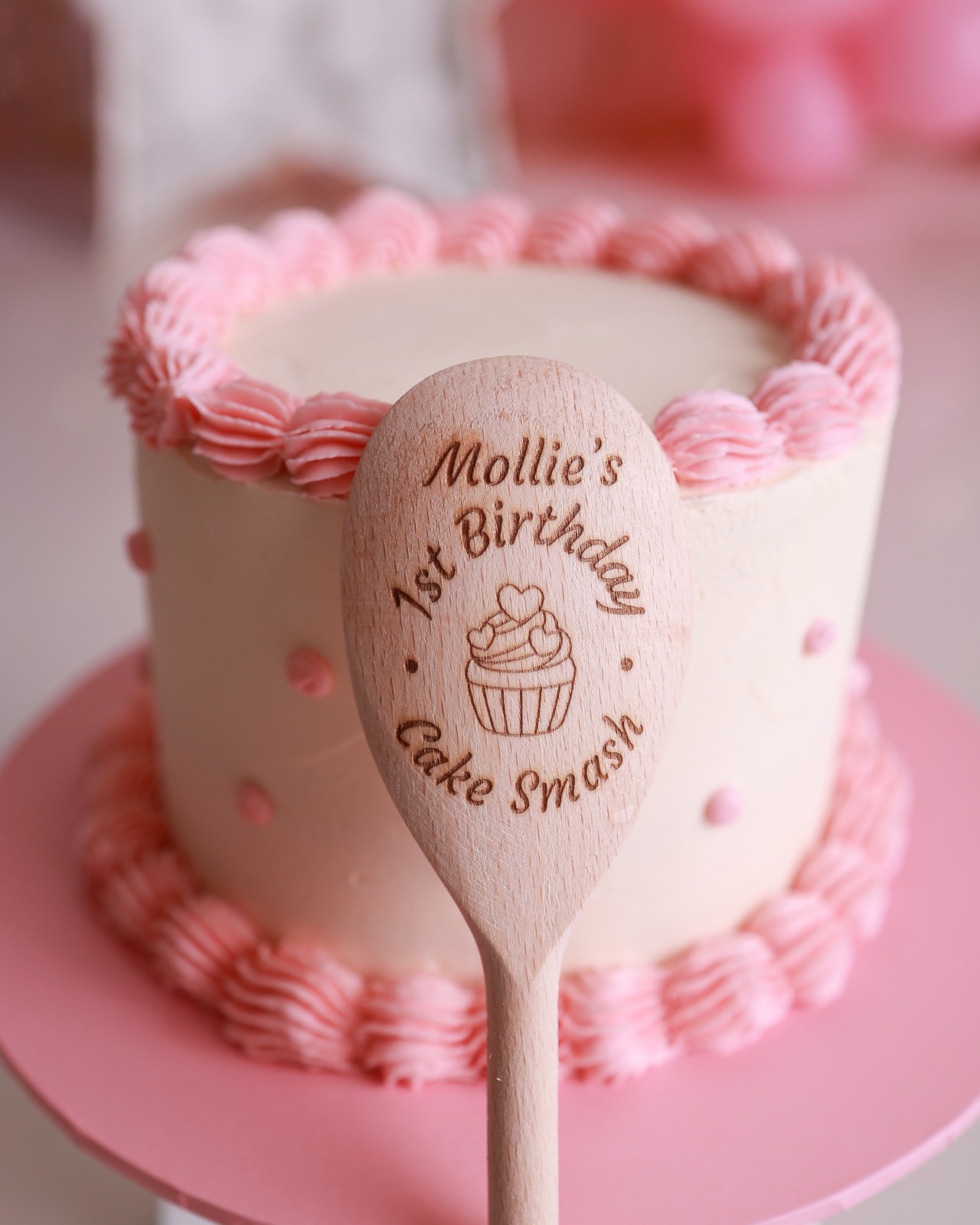 A personalised wooden cake-smash spoon propped against a cream birthday cake with pink piping