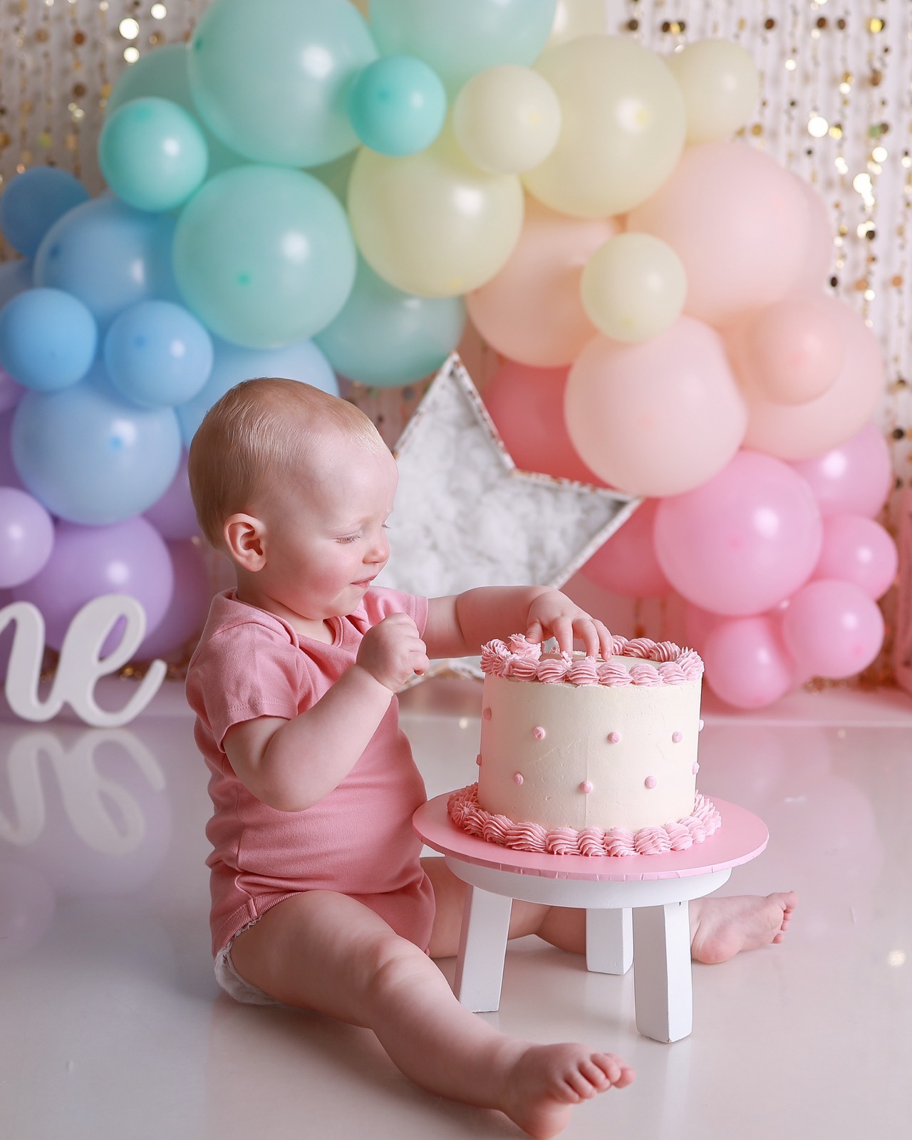 Baby girl in a pink outfit reaching for a cream birthday cake with pink piping and dots, pastel balloon backdrop