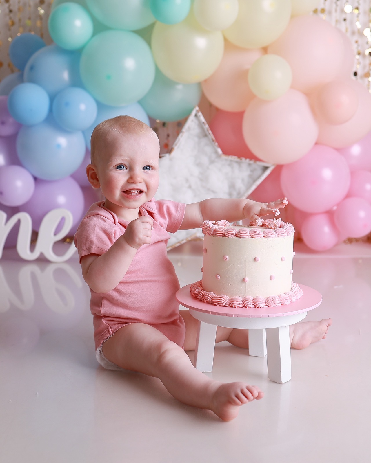 Baby girl sitting beside a cream birthday cake with pink piping, hand raised, pastel balloon backdrop