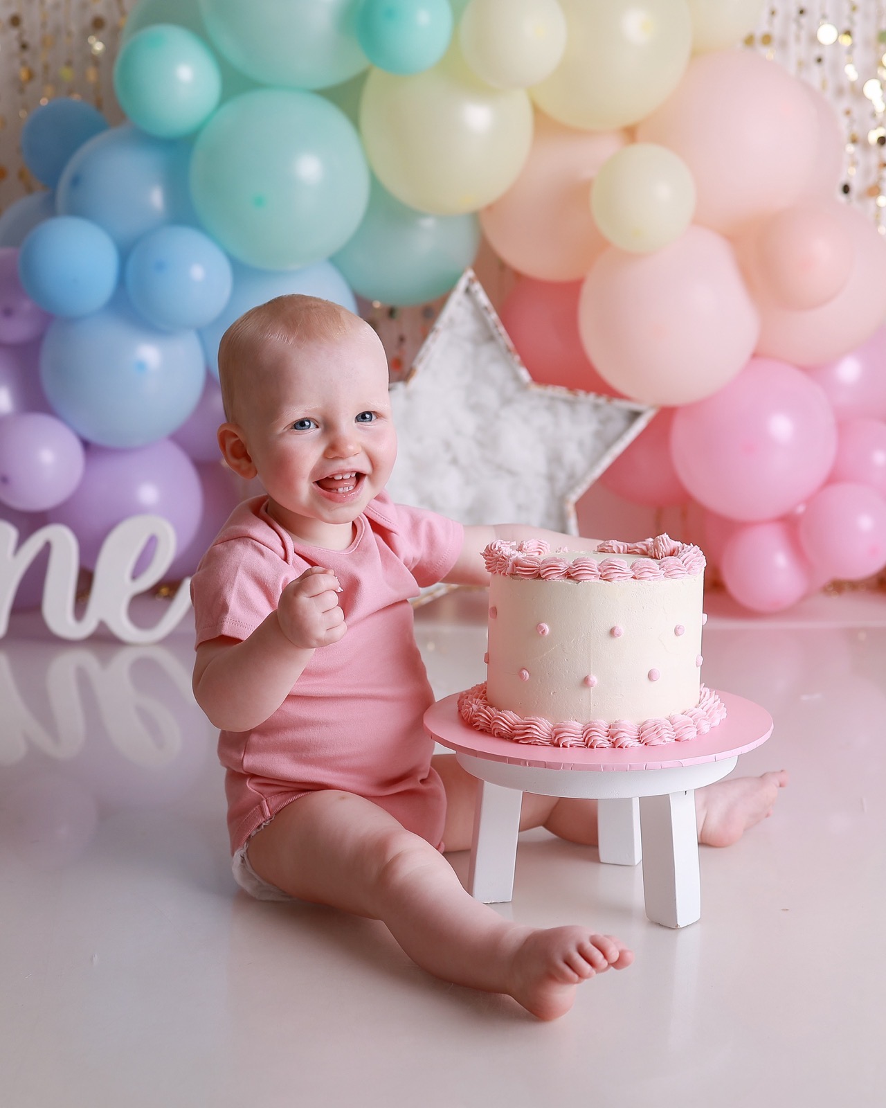 Baby girl smiling widely with her hand resting on a cream birthday cake, pastel balloon backdrop