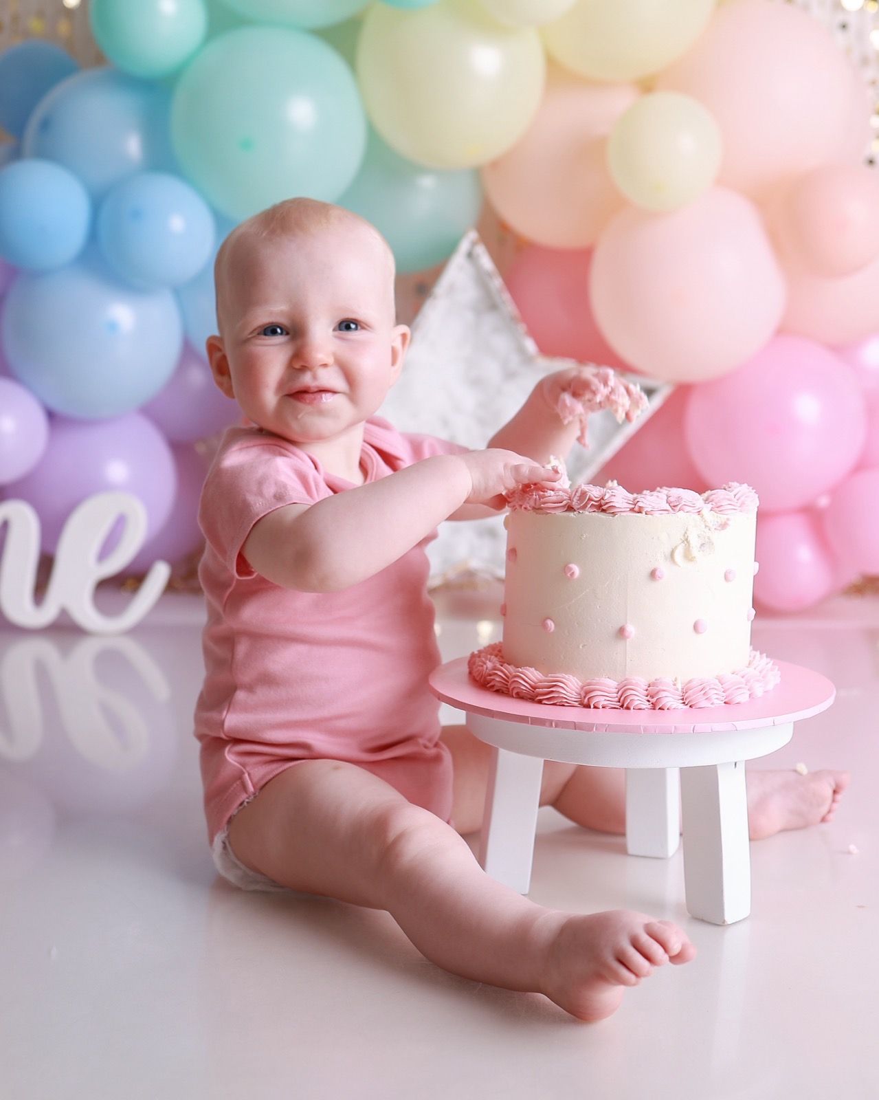Baby girl with icing on her fingers beside a cream birthday cake, pastel balloon backdrop