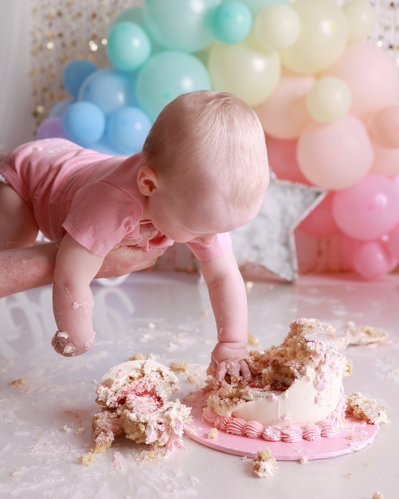 Baby girl leaning forward over a completely smashed cake on the floor, supported by an adult's arm
