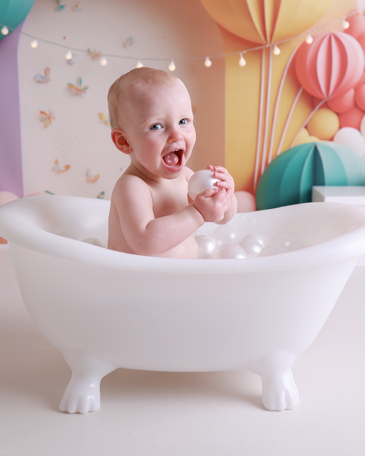 Toddler boy laughing in a white bath tub holding a bath ball, colourful hot-air-balloon-themed backdrop