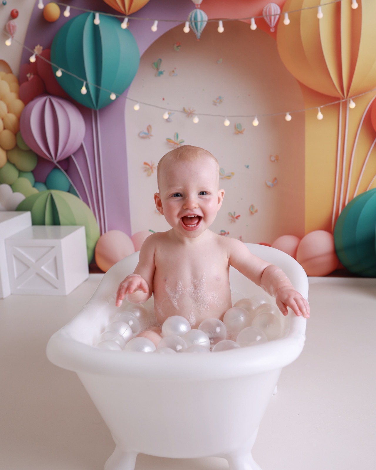 Toddler boy smiling in a white bath tub with pearl bath balls, colourful hot-air-balloon-themed backdrop