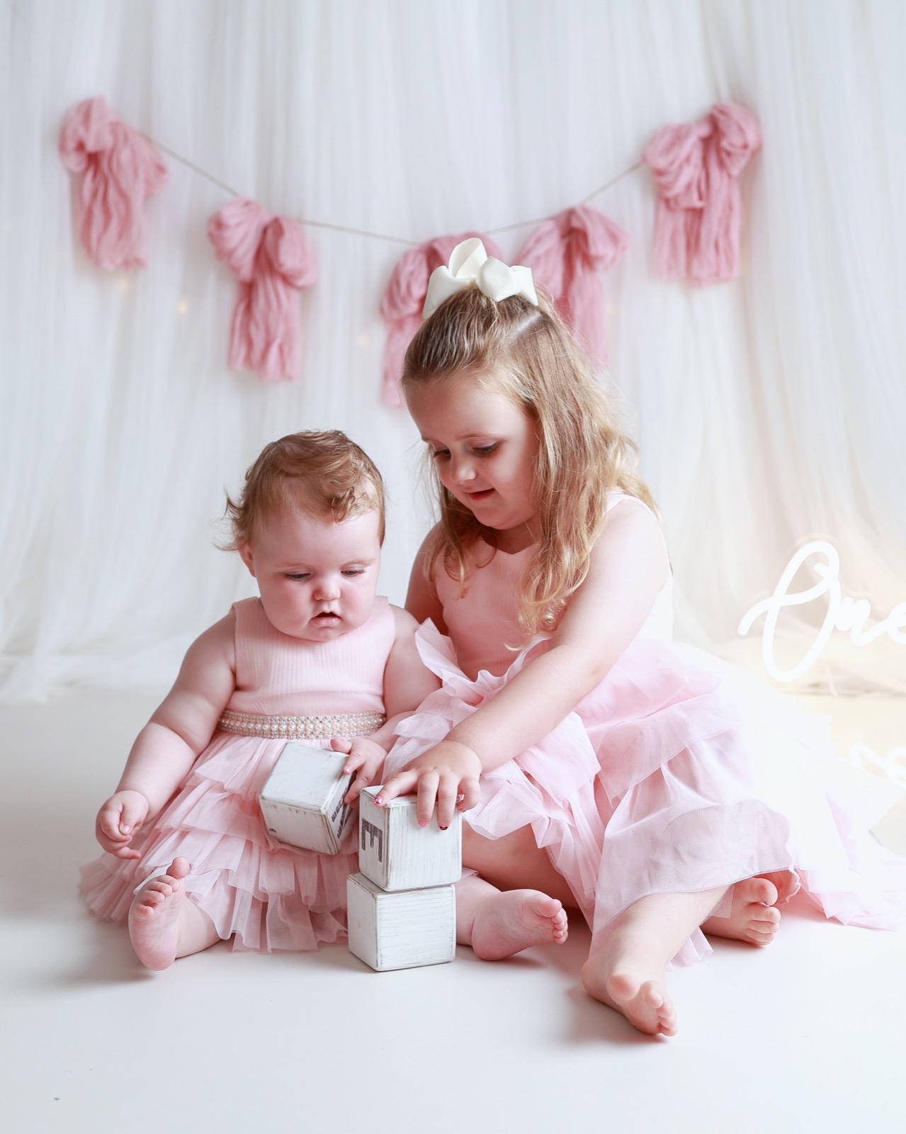 Baby girl and her older sister playing with wooden alphabet blocks, pink tassel garland backdrop