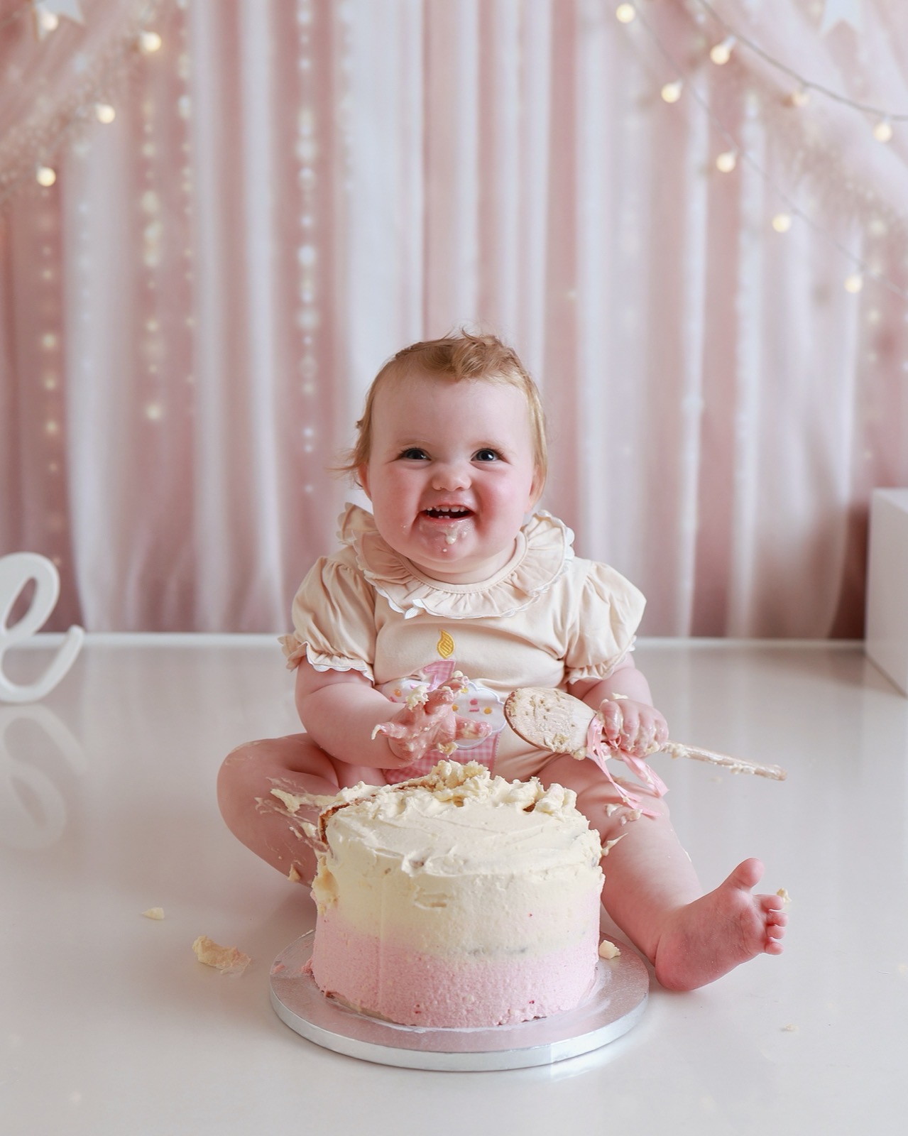 Baby in a cream outfit laughing with cake on her face, holding a wooden spoon, pink fairy-light curtain backdrop