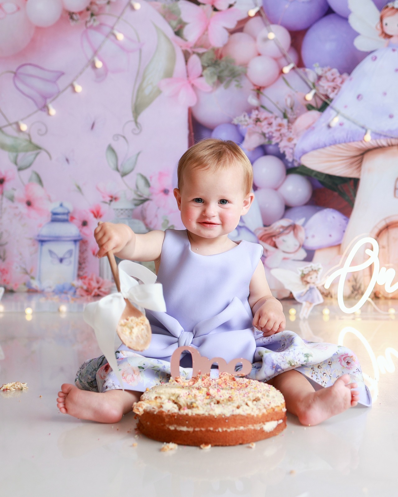 Toddler girl in a lilac bow dress holding a wooden spoon beside a sprinkle-topped sponge cake, whimsical fairy and mushroom backdrop