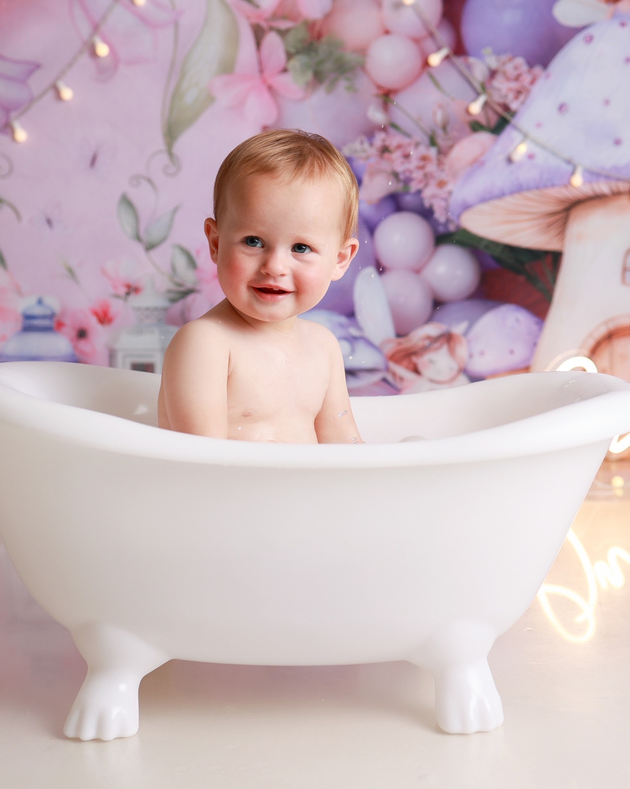Toddler girl smiling in a white bath tub, whimsical fairy and mushroom floral backdrop