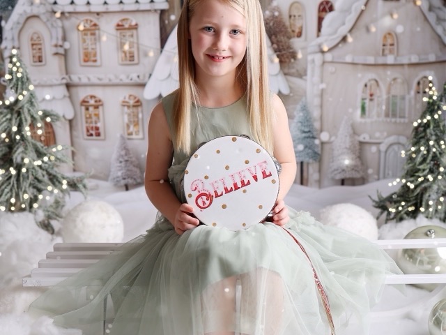 Girl smiling in front of a gingerbread village backdrop, holding a &ldquo;Believe&rdquo; tambourine, from a Christmas mini session