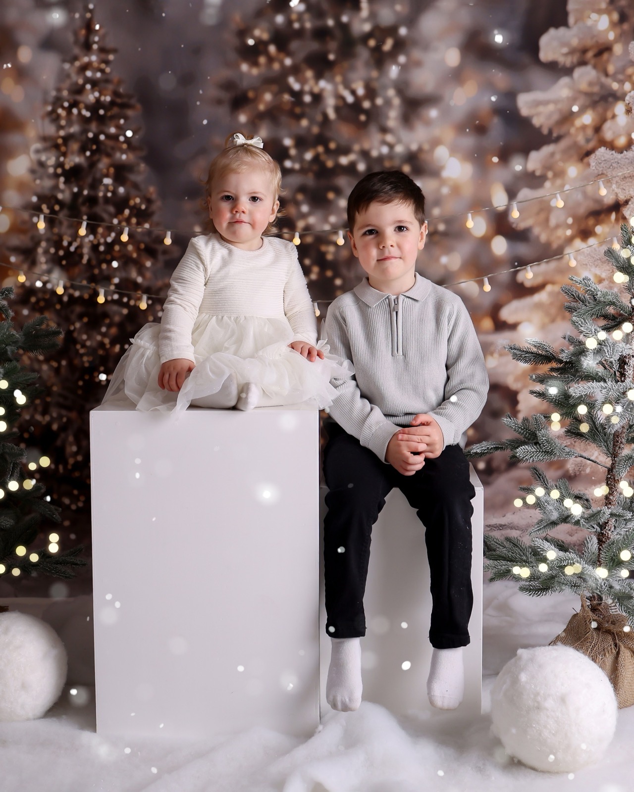 Toddler girl and her older brother sitting together on a white plinth, both in white outfits, snowy Christmas backdrop