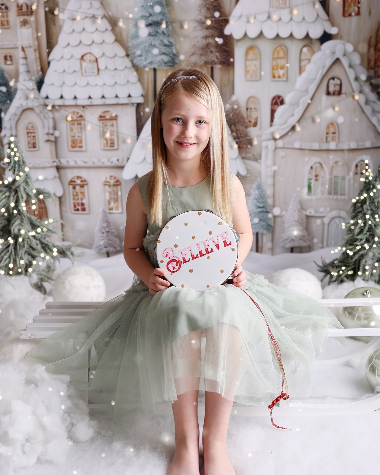 Girl in a sage green tulle dress holding a tambourine that reads 'Believe', snowy gingerbread village backdrop
