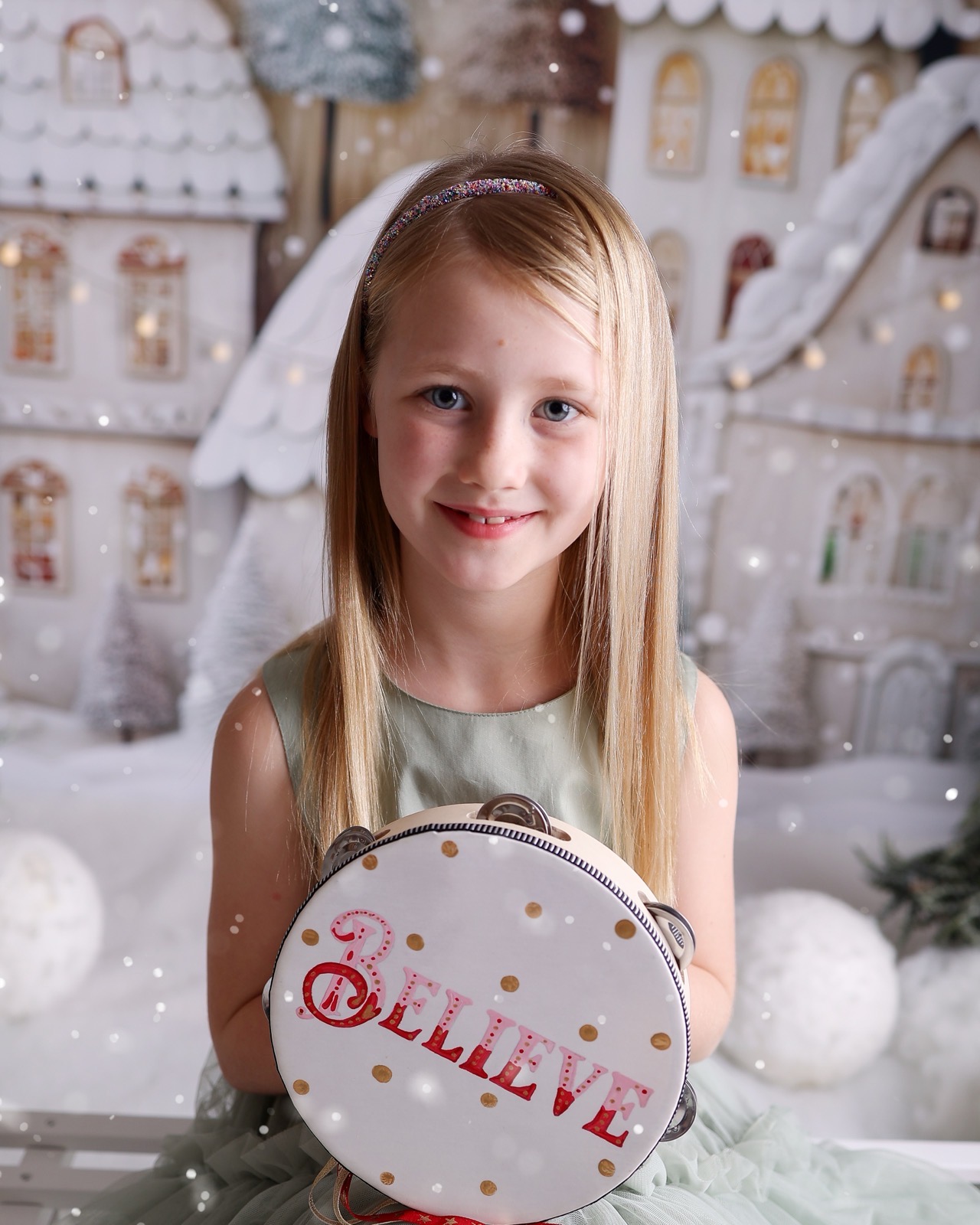 Close-up portrait of a girl in a sage green dress holding a 'Believe' tambourine, snowy village backdrop
