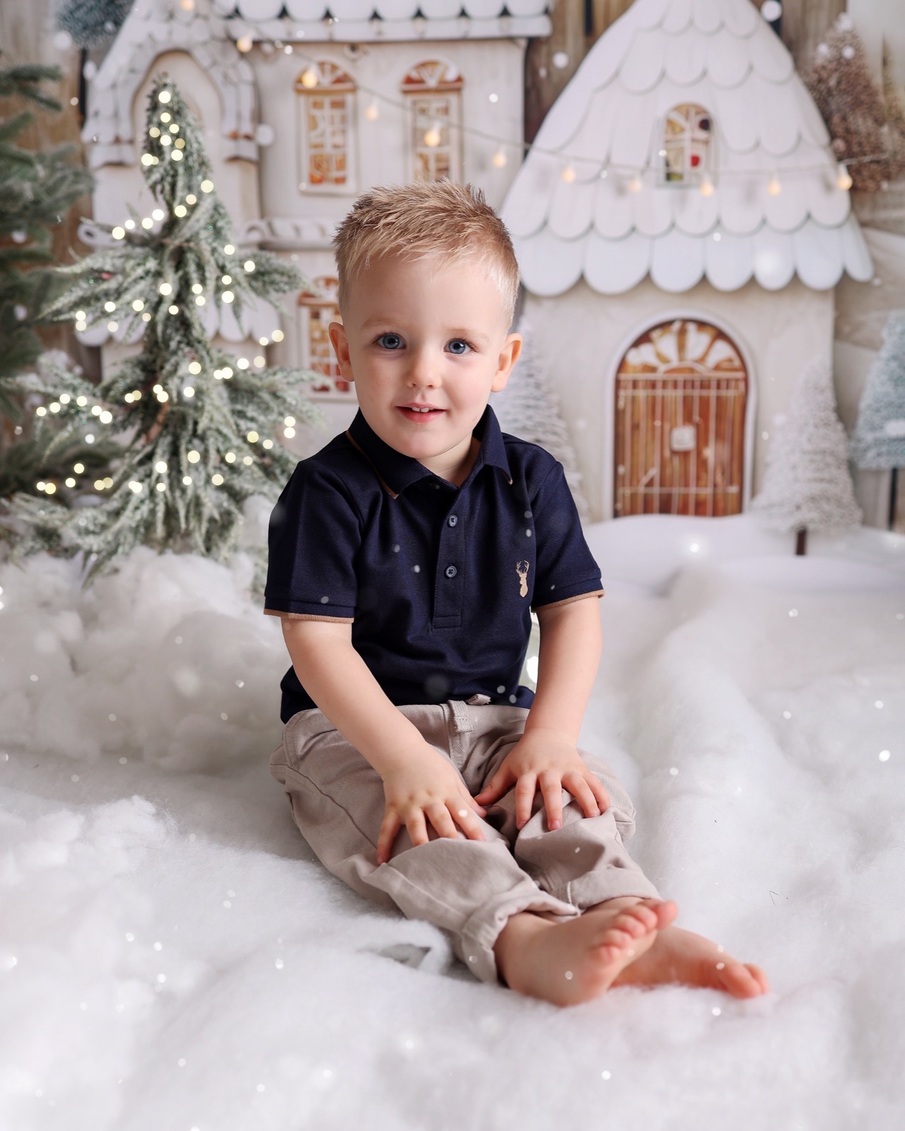 Toddler boy sitting barefoot in fake snow, navy polo shirt with an embroidered stag, gingerbread house backdrop