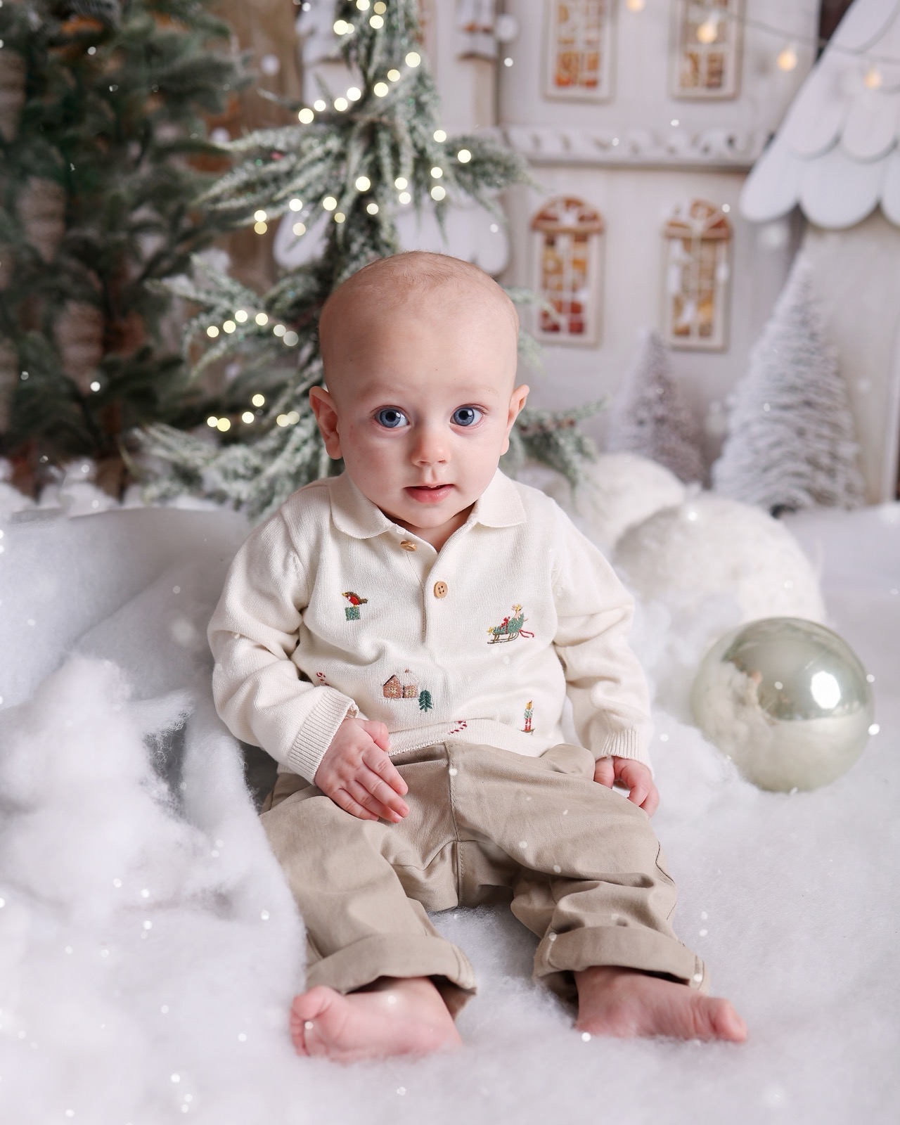 Baby boy sitting in fake snow wearing a cream jumper with embroidered Christmas motifs