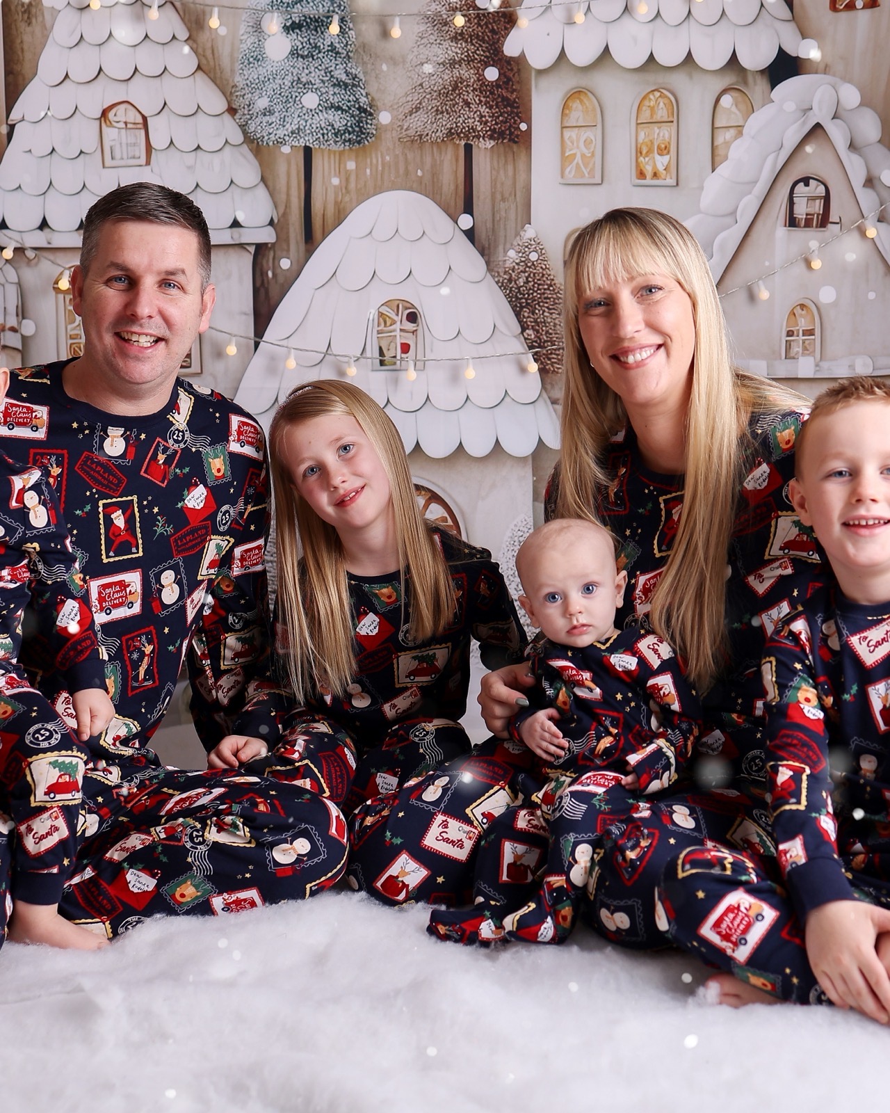 Family of five in matching Christmas pajamas printed with Santa and reindeer, sitting together on fake snow