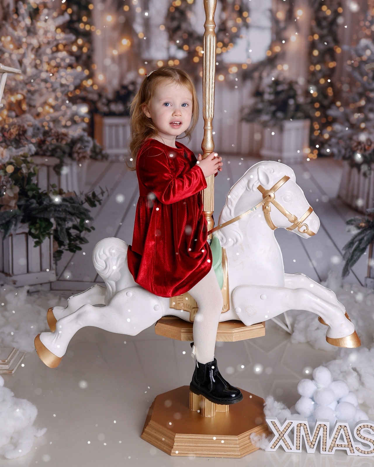 Toddler girl in a red velvet dress riding a white and gold carousel horse, an 'XMAS' sign nearby