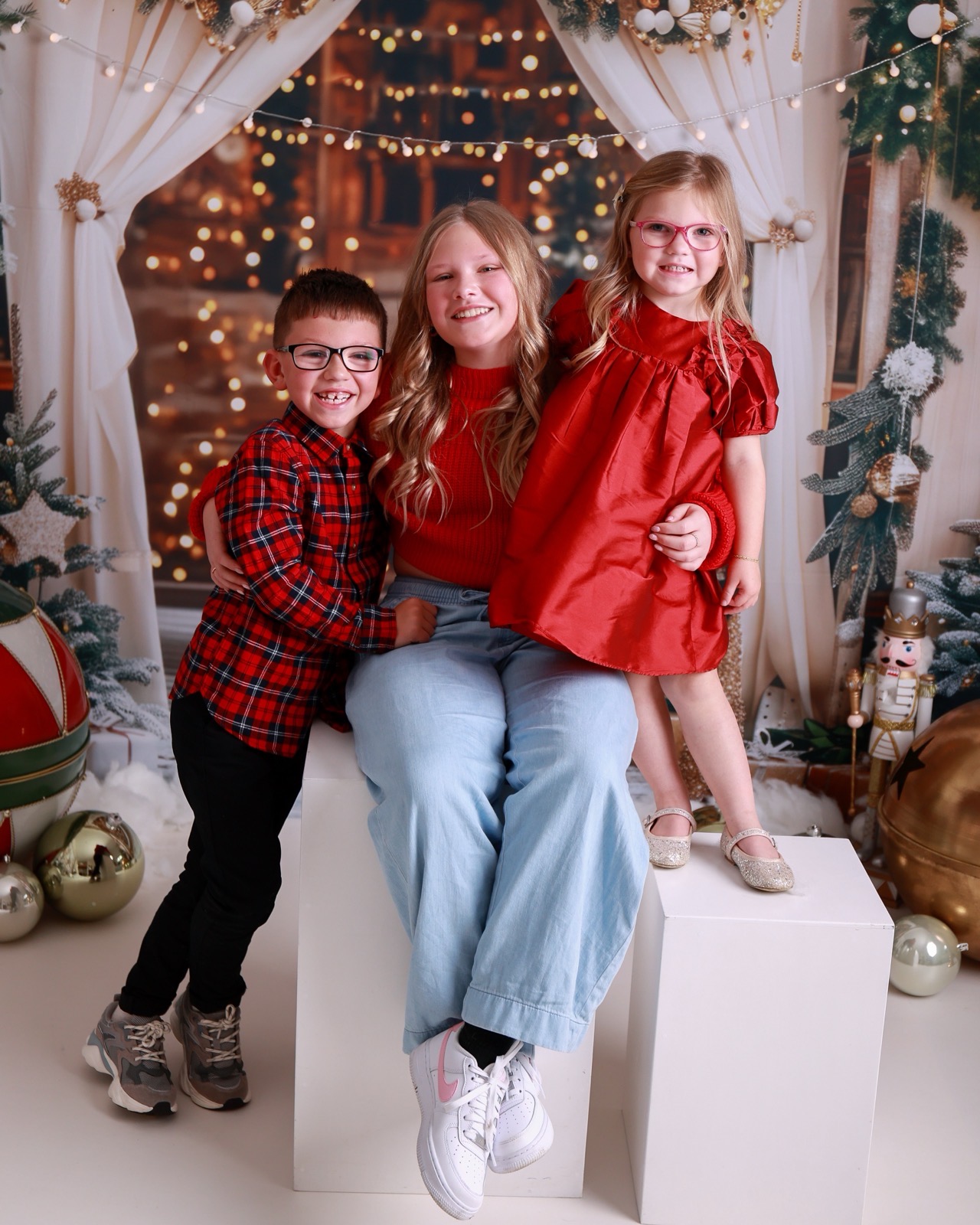 Three siblings sitting together on white plinths, boy in a checked shirt and two girls in red, fairy-light curtain backdrop