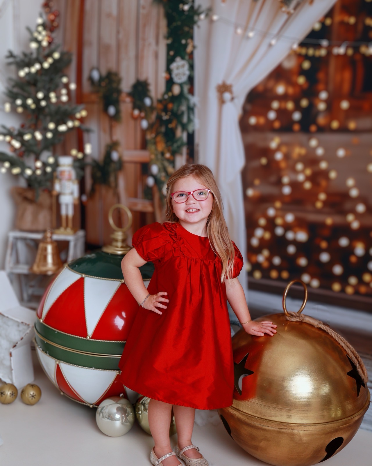 Girl in a red dress and glasses standing beside a giant red, white and gold bauble and jingle bell props