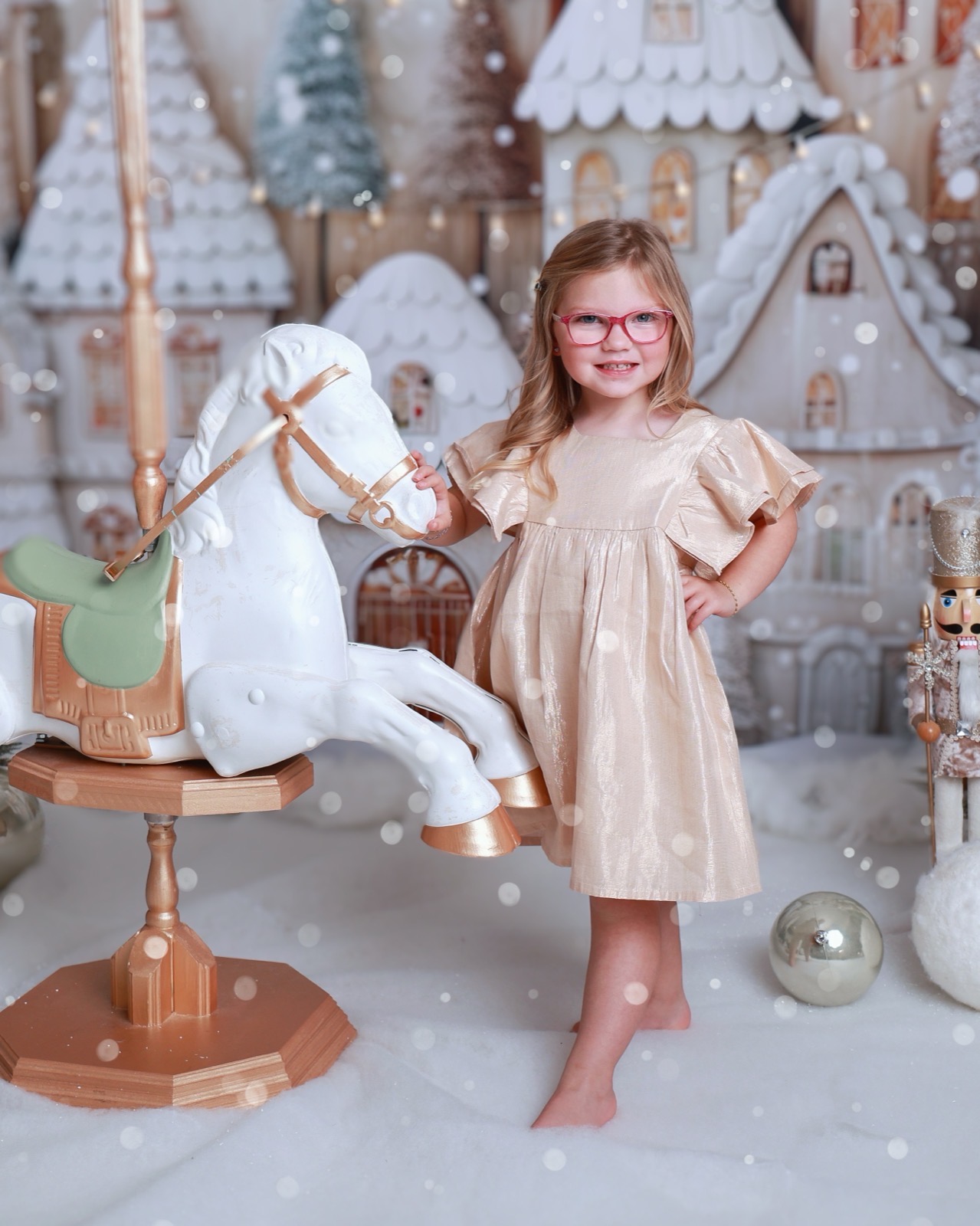 Girl in a gold dress standing beside a white and gold carousel horse, gingerbread village backdrop
