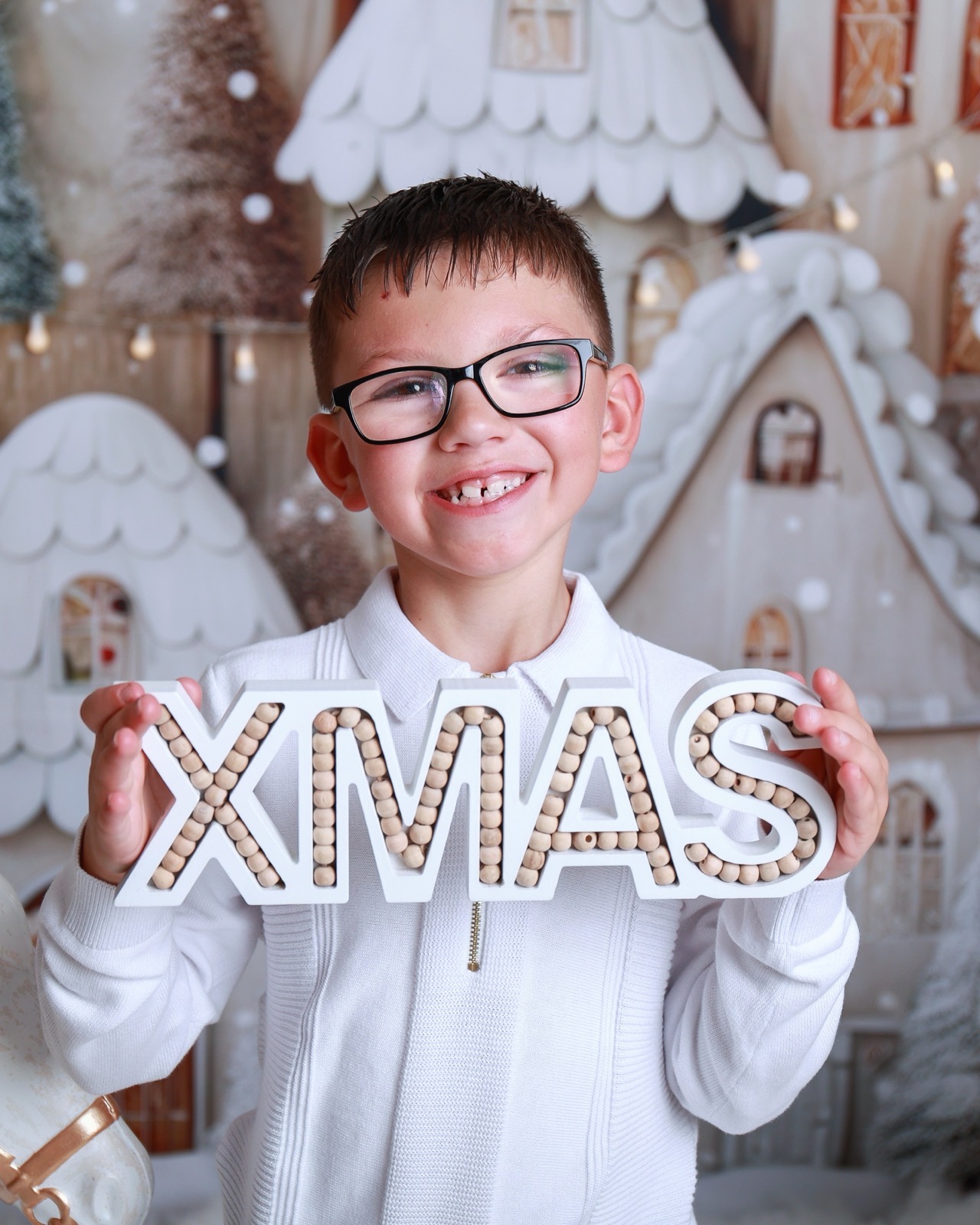 Boy in glasses holding a large wooden 'XMAS' sign, gingerbread village backdrop