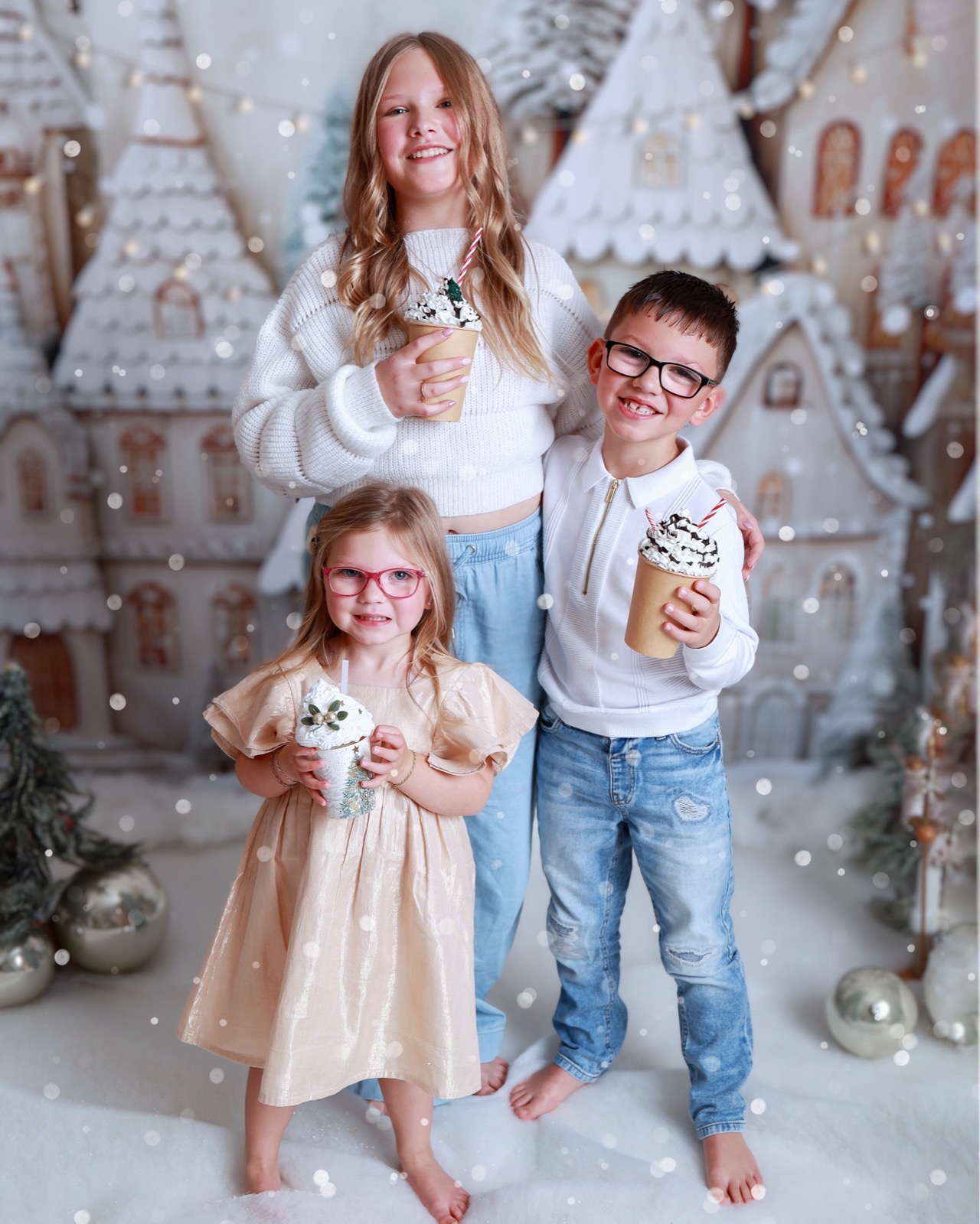 Three siblings holding decorated hot chocolate cups topped with cream, gingerbread village backdrop