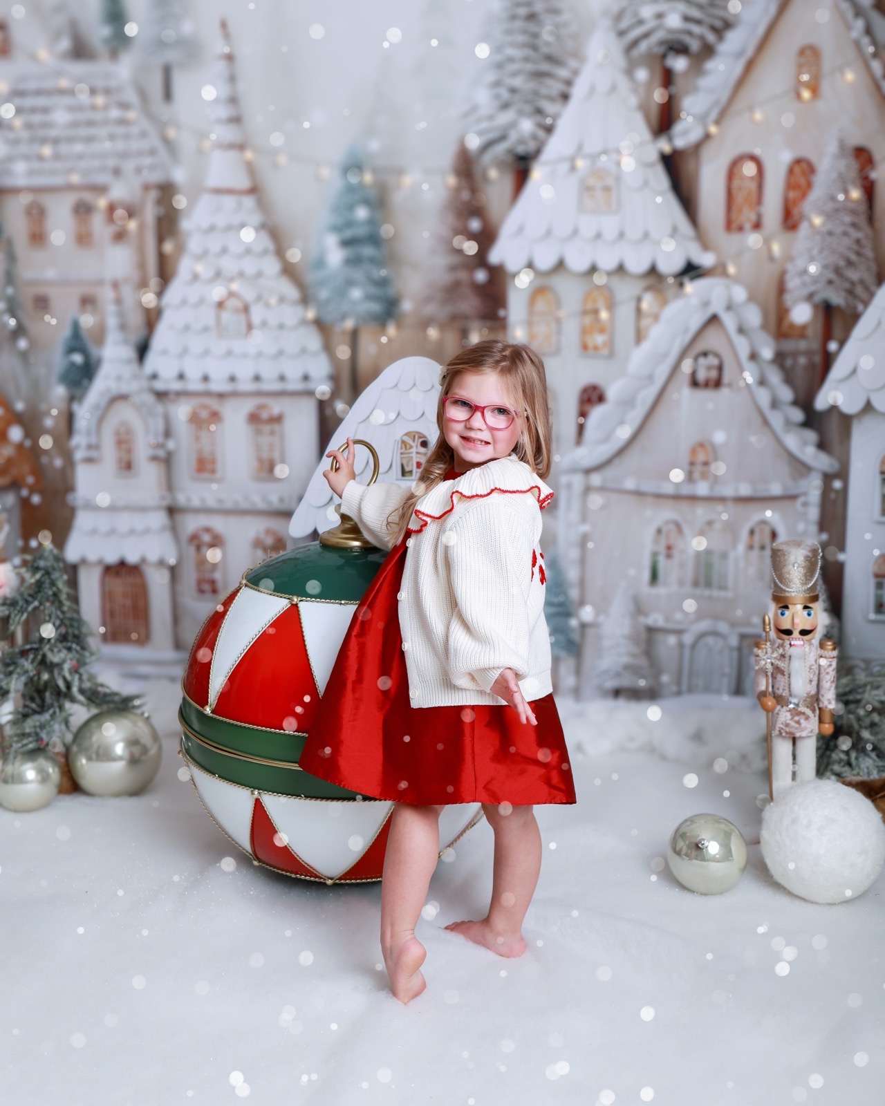 Girl in a cream cardigan and red skirt leaning on a giant ornament, gingerbread village backdrop